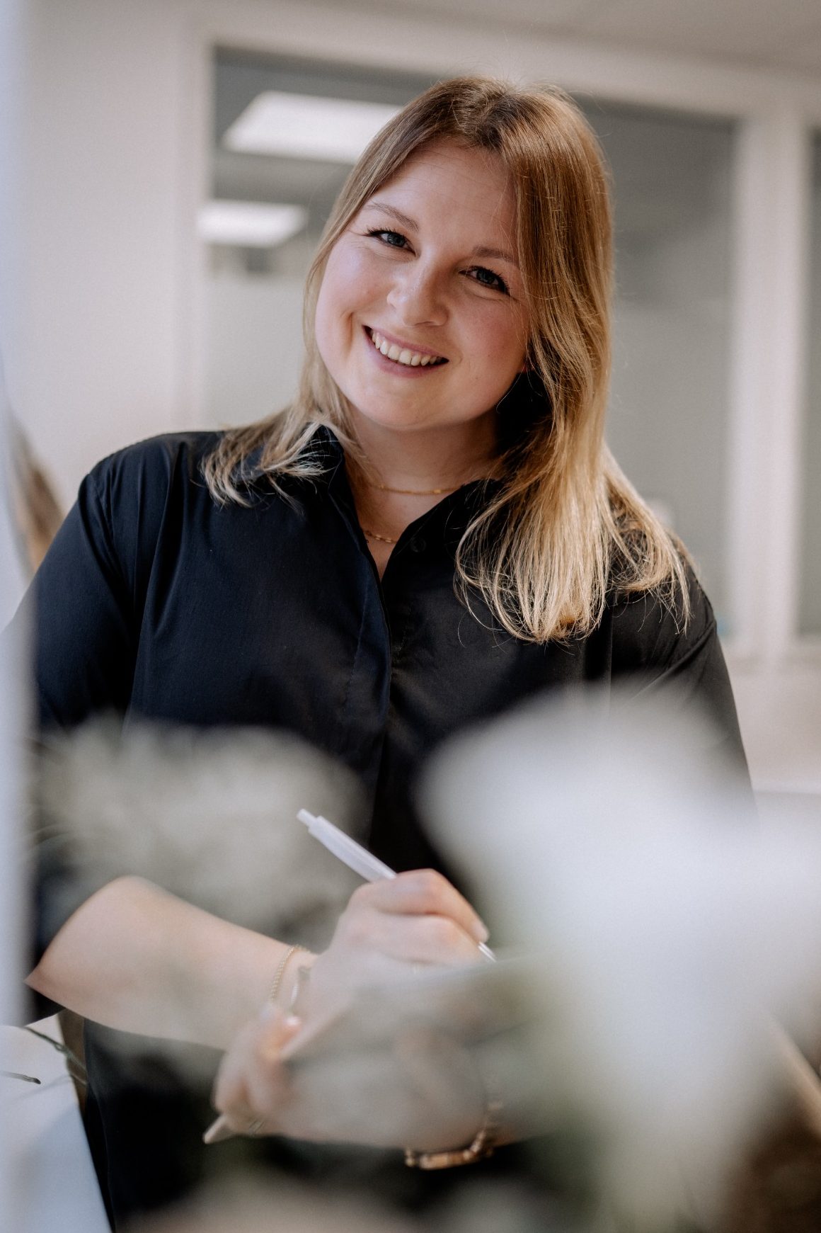 Lisa Lamparter Lisa Lamparter mit schwarzer Bluse und Kugelschreiber in der Hand lehnt an der Wand und lächelt.